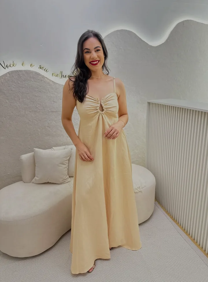 Modelo com vestido longo bege de alças e recorte no busto, diante de sofá curvo